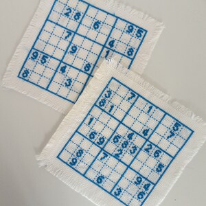 May include: Two white fabric squares with blue embroidery of a 9x9 grid with numbers in each square. The numbers are 95, 286, 48, 7, 6, 954, 8, 8, 57, 3, 81, 26, 683, 69, 7, 190, 46, 6.