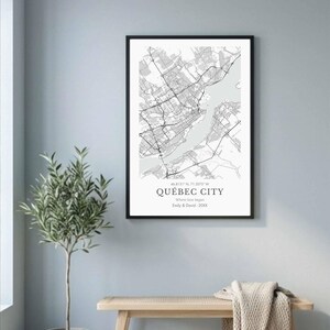 May include: A framed black and white map print of Québec City, Canada, with street and river details. The print includes the city's name, coordinates, and a personalized message. The artwork hangs on a light blue wall, with a bench and plant in the foreground.