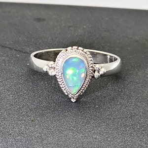 Può includere: Un anello d'argento con una pietra di opale a forma di pera incastonata in una montatura d'argento dettagliata. L'anello ha una fascia delicata e un design decorativo attorno alla pietra.