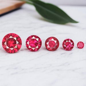 May include: Five round, faceted, red gemstones of varying sizes are arranged in a row. The gems have a starburst pattern in the center. A green leaf and a wooden surface are in the background.