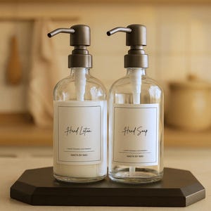May include: Two clear glass pump bottles with matte dark gray dispensers. One bottle contains white hand lotion, the other is empty for hand soap. Each features a white rectangular label with black text reading Hand Lotion, Hand Soap, and NIKITA BY NIKI.