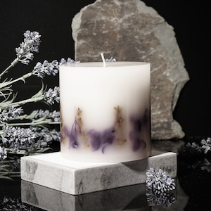 Könnte beinhalten: Eine weiße Säulenkerze mit lila und goldenen floralen Akzenten, die auf einem grauen Marmorblock ruht. Lavendelzweige und ein großer Stein sind vor einem schwarzen Hintergrund angeordnet. Die Kerze ist unparfümiert.