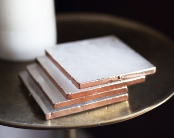 May include: Set of four white square coasters with a copper trim. The coasters are stacked on top of each other.