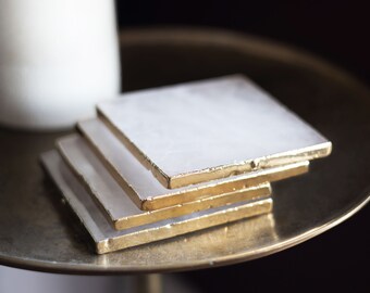 May include: Four white square coasters with gold trim stacked on top of each other. The coasters are on a gold metal tray.