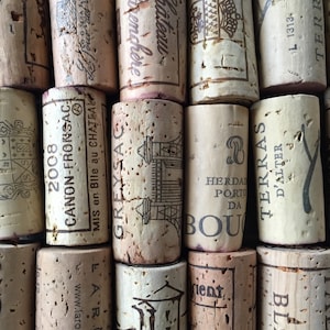 May include: A close-up shot of used wine corks. The corks are cylindrical and made of natural cork. Various corks have printed text and designs, including winery names and dates. The corks are arranged closely together, showing their varied markings.