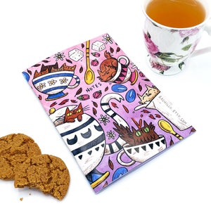 May include: A pink notebook with a whimsical illustration of cats in teacups. The illustration features a variety of cats in different poses, with some holding spoons and others sipping from teacups. The notebook has the word "NOTES" printed on the front cover.