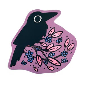 May include: A black crow illustration perched on a branch with pink leaves and blue flowers. The crow has a white eye and is set against a purple background. The artwork is a sticker.