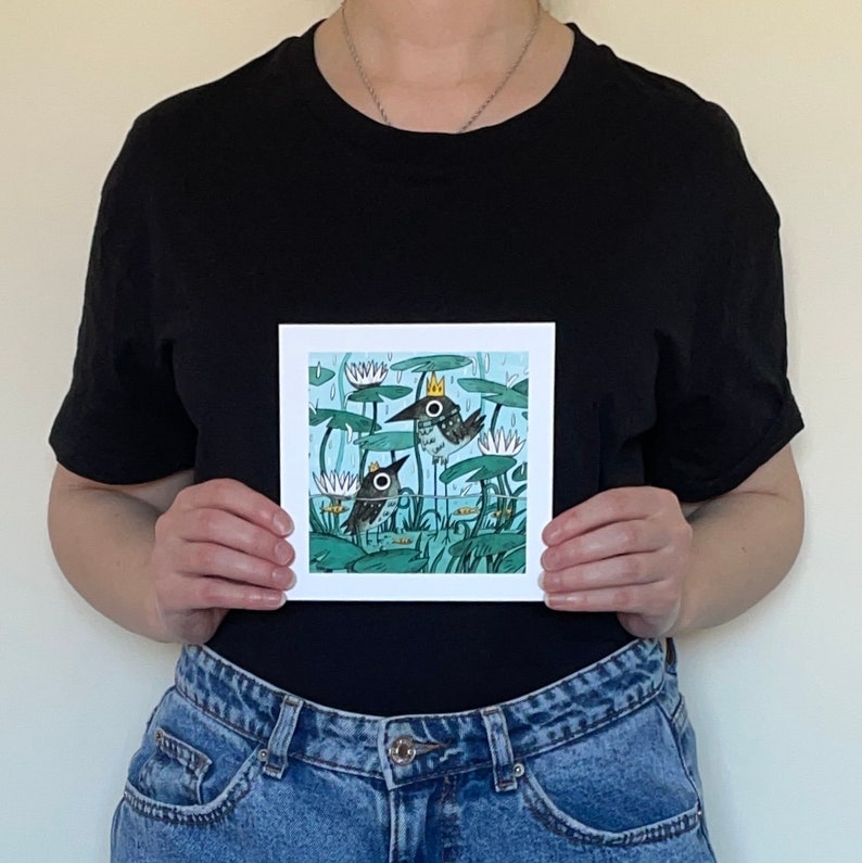 May include: Square art print featuring two cartoon birds with crowns, set in a pond with lily pads and rain. The birds are black and white with yellow crowns. The background is a light blue with white rain lines.