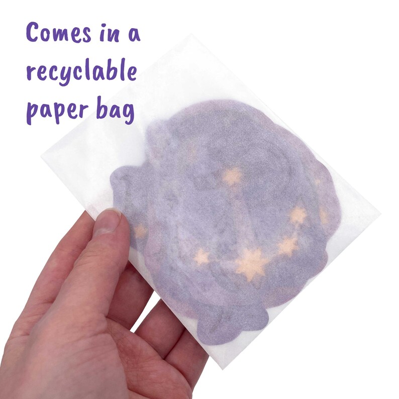 May include: A purple and gold celestial-themed item is held in a recyclable paper bag. The item has a star pattern and is visible through the translucent paper. The text "Comes in a recyclable paper bag" is in purple.
