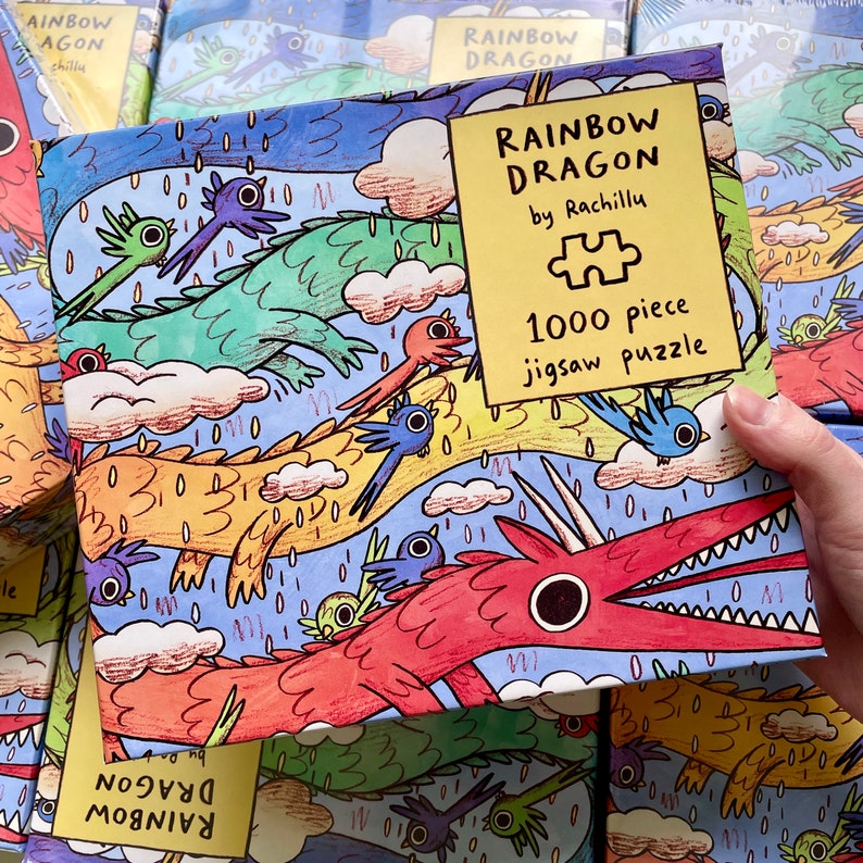 May include: A 1000 piece jigsaw puzzle box titled Rainbow Dragon by Rachillu. The box features a colorful illustration of red, green, and yellow dragons flying among blue birds and pink clouds against a light blue background with rain.