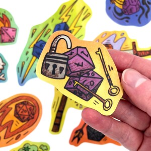 May include: A collection of colorful, illustrated stickers with a fantasy theme. One sticker features a padlock, purple dice, and keys. Other stickers show a lightning bolt, a fireball, and other dice. The stickers are held by a hand.