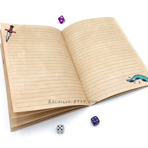 May include: Open brown notebook with lined pages, featuring a red and black dagger illustration on the left page and a green and blue sea serpent illustration on the right page.  The notebook is surrounded by blue dice.