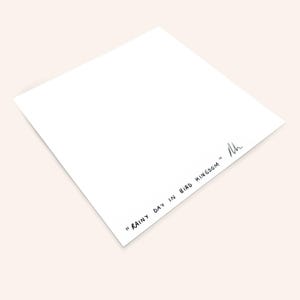 May include: A white, square art print with the text "RAINY DAY IN BIRD KINGDOM" in black script. The print is angled, showing a corner and the text. A signature is present.