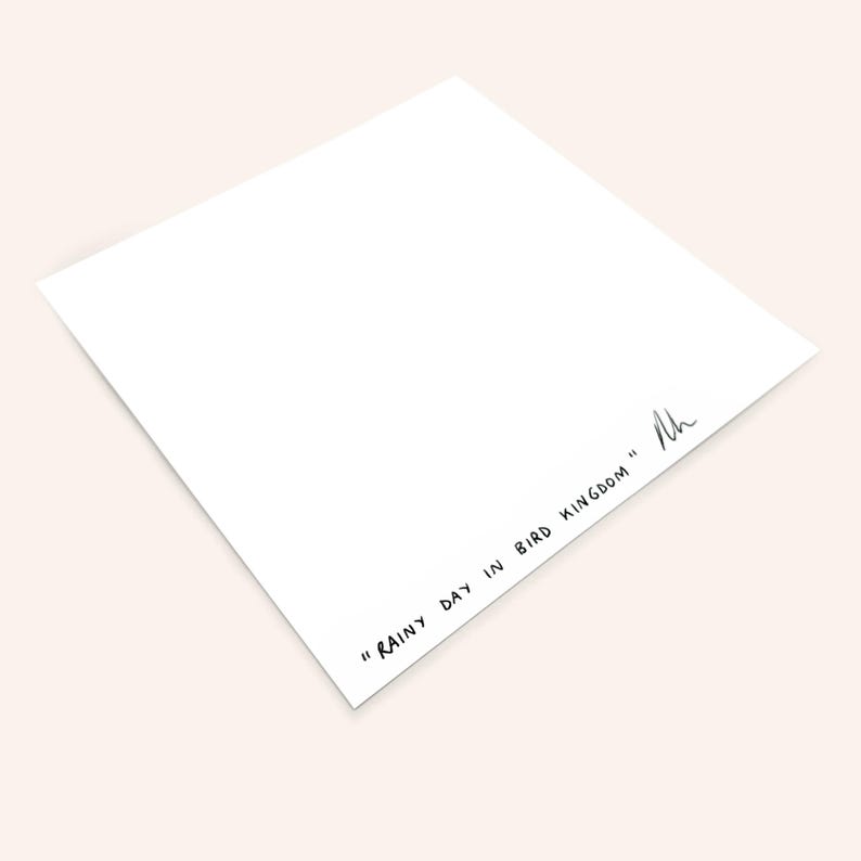 May include: A white, square art print with the text "RAINY DAY IN BIRD KINGDOM" in black script. The print is angled, showing a corner and the text. A signature is present.
