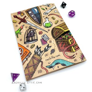 May include: A brown notebook with a colorful illustration of a treasure chest, a sword, a bird in a cage, a potion bottle, and other fantasy-themed items. The notebook has the text "rachillu.com" and "RACHILLU. ETSY. COM" printed on it.