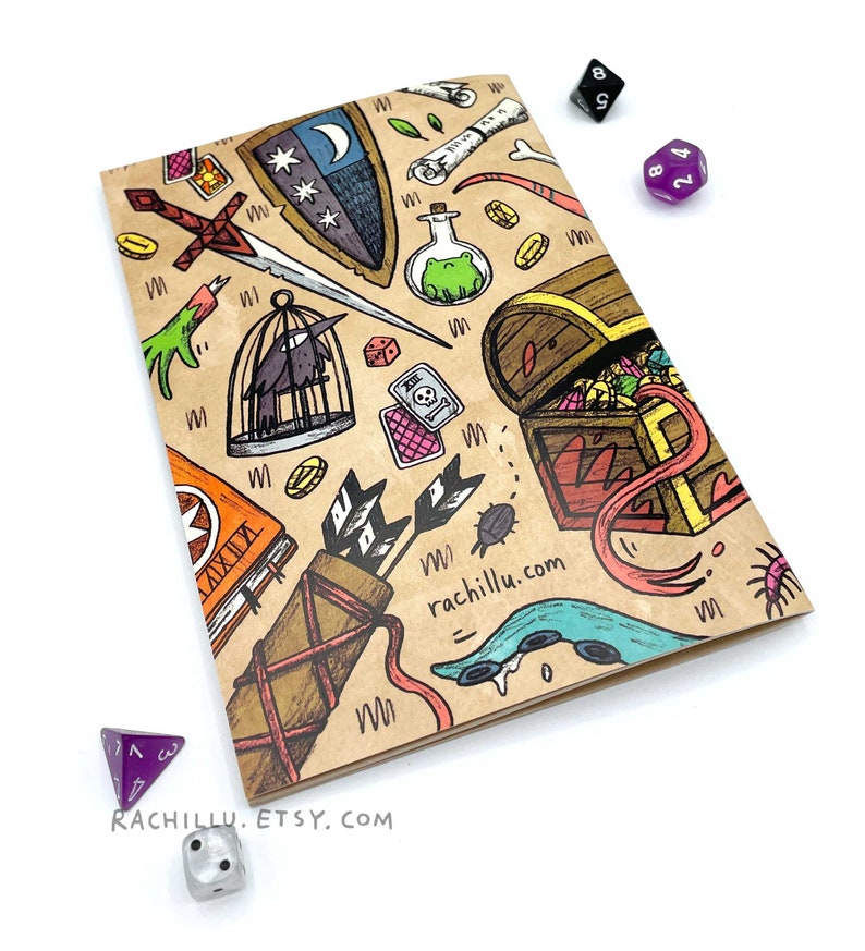 May include: A brown notebook with a colorful illustration of a treasure chest, a sword, a bird in a cage, a potion bottle, and other fantasy-themed items. The notebook has the text "rachillu.com" and "RACHILLU. ETSY. COM" printed on it.