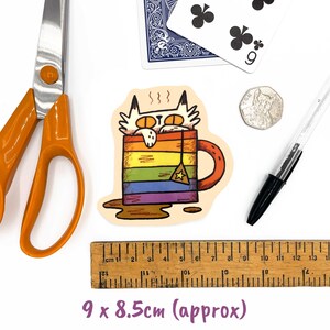Pu&ograve; includere: Un adesivo raffigurante un gatto in una tazza arcobaleno con una bustina di t&egrave;. L'adesivo misura circa 9 x 8,5 cm. Altri elementi includono forbici, una carta da gioco, una moneta e una penna.