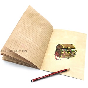 May include: An open lined journal with a brown cover. The right page has a hand-drawn illustration of a treasure chest overflowing with jewels and coins. The text "rachillu.etsy.com" is printed on the left page. A red pencil is lying on the open journal.