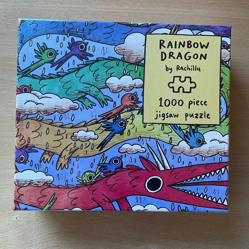 May include: A 1000 piece jigsaw puzzle box titled Rainbow Dragon by Rachillu. The box features a colorful illustration of multiple dragons and birds flying through a blue sky with clouds and rain. The text on the box reads Rainbow Dragon by Rachillu 1000 piece jigsaw puzzle.