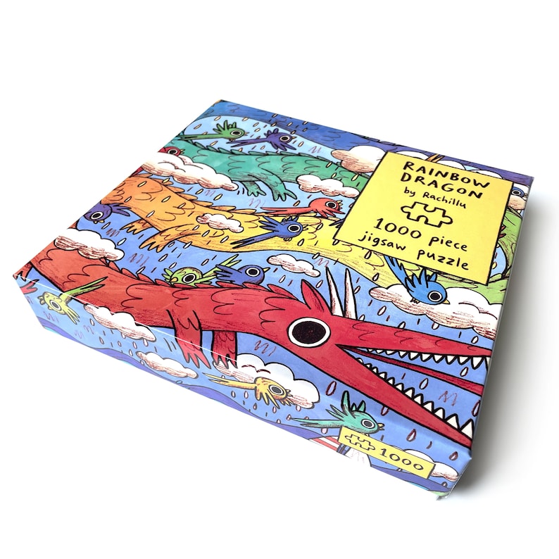 May include: A 1000 piece jigsaw puzzle box titled Rainbow Dragon by Rachillu. The box features a colorful illustration of long, multi-colored dragons flying through a blue sky with clouds, rain, and small birds. The text is clearly visible.