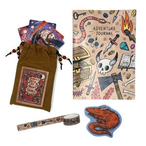 May include: A brown fabric drawstring pouch labelled Deck of Many Things, a notebook titled Adventure Journal with fantasy illustrations, a roll of patterned washi tape, and a die-cut sticker of a red dragon, all featuring colourful cartoon art.