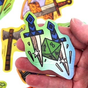 May include: A sticker featuring two crossed swords with blue handles and a green die. The sticker has a green and blue gradient background with white stars and is held in a hand. Other stickers with similar designs are in the background.