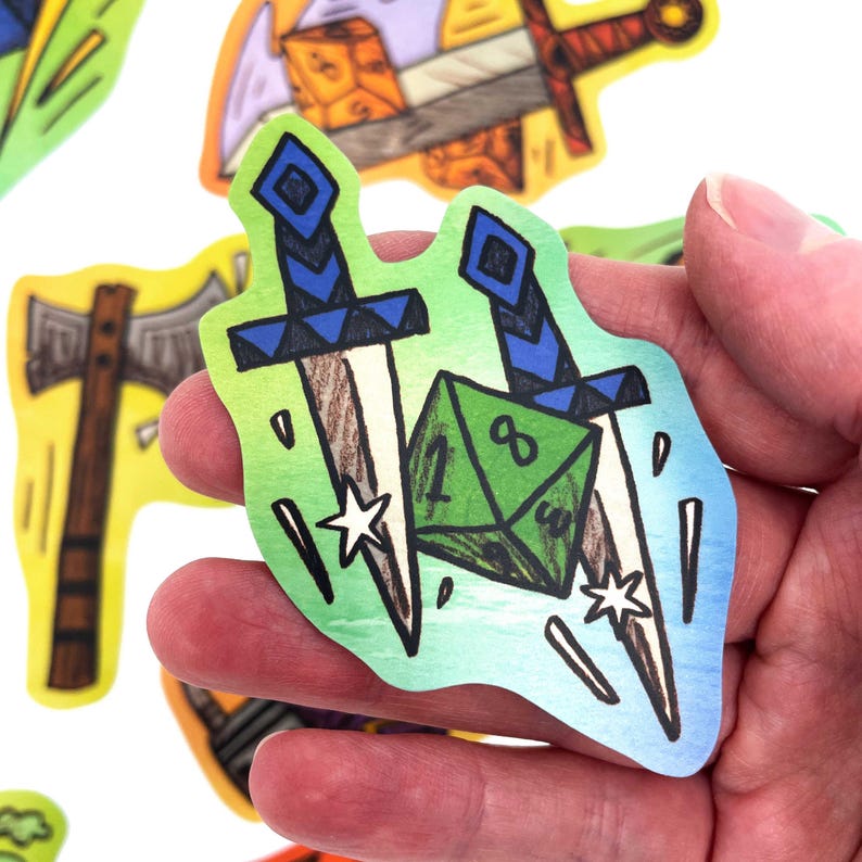May include: A sticker featuring two crossed swords with blue handles and a green die. The sticker has a green and blue gradient background with white stars and is held in a hand. Other stickers with similar designs are in the background.