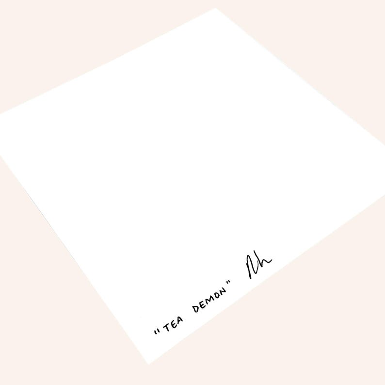 Pu&ograve; includere: Un foglio di carta bianco con la scritta "TEA DEMON" in nero, con un carattere scritto a mano. La carta &egrave; angolata, con una firma nell'angolo. Lo sfondo &egrave; di colore pesca chiaro.