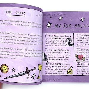 May include: An open book with purple pages and illustrations of tarot cards. The book features the Major Arcana cards, including The Fool, The High Priestess, The Magician, The Empress, and The Hierophant. The text on the page reads "The Cards" and "Major Arcana".