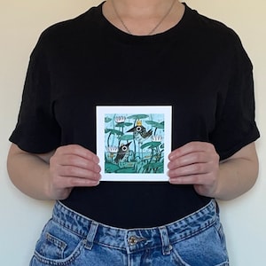 May include: Square art print featuring a whimsical illustration of two birds in a pond with lily pads and water lilies. One bird wears a crown. The artwork is set against a light blue background with green and white accents.