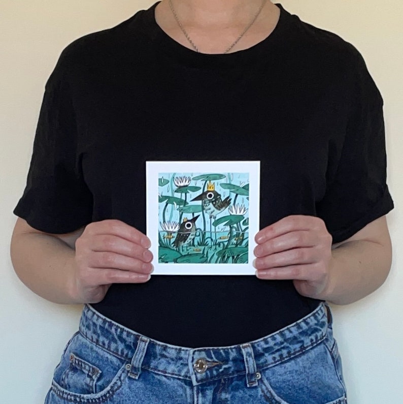 May include: Square art print featuring a whimsical illustration of two birds in a pond with lily pads and water lilies. One bird wears a crown. The artwork is set against a light blue background with green and white accents.