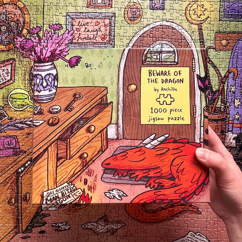 May include: A 1000-piece jigsaw puzzle box with a cartoon dragon illustration. The box reads "BEWARE OF THE DRAGON" and "1000 piece jigsaw puzzle". The puzzle is set in a room with a wooden door and furniture.