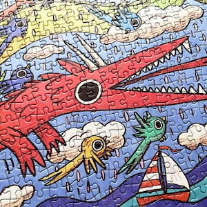 May include: A jigsaw puzzle featuring a large red dragon with sharp teeth, a yellow creature, a green creature, and a small sailboat with red and white stripes on a blue sea. The scene includes white clouds and rain against a blue background.