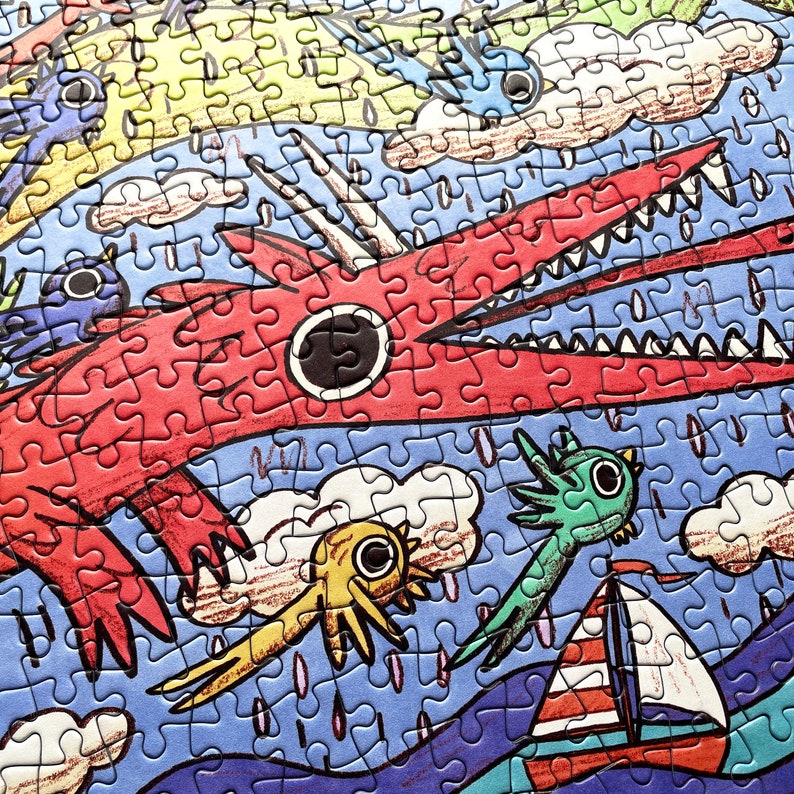 May include: A jigsaw puzzle featuring a large red dragon with sharp teeth, a yellow creature, a green creature, and a small sailboat with red and white stripes on a blue sea. The scene includes white clouds and rain against a blue background.