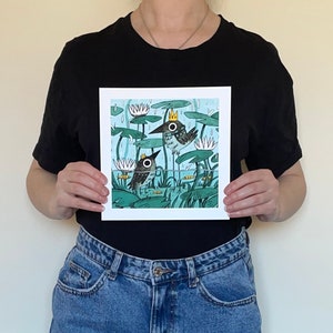 May include: Square art print featuring a whimsical illustration of two birds in a pond with lily pads and water lilies. The birds wear crowns and are set against a light blue background with rain. The artwork has a cartoon style.