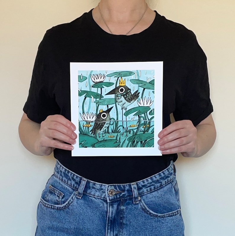 May include: Square art print featuring a whimsical illustration of two birds in a pond with lily pads and water lilies. The birds wear crowns and are set against a light blue background with rain. The artwork has a cartoon style.