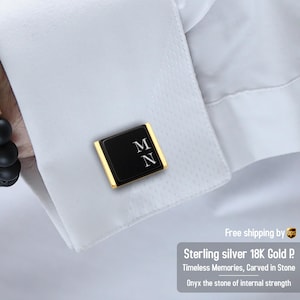 Op de afbeelding: Zwarte en gouden manchetknopen met de initialen "MN" in wit. De manchetknopen zijn rechthoekig met een zwart midden en gouden rand. De afbeelding bevat tekst: "Sterling silver 18K Gold P" en "Onyx the stone of internal strength."