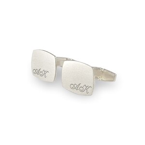 May include: A pair of silver cufflinks with a square shape and a brushed finish. The cufflinks are engraved with the initials "A.K."
