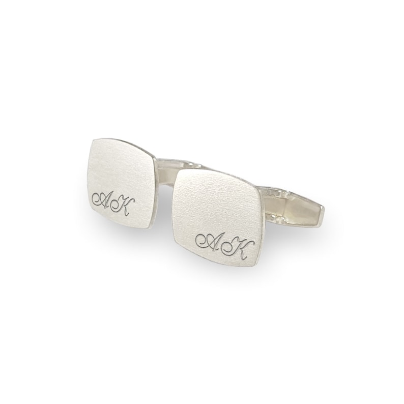 May include: A pair of silver cufflinks with a square shape and a brushed finish. The cufflinks are engraved with the initials "A.K."