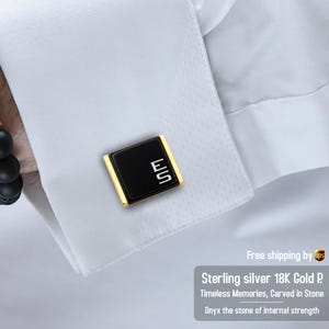 May include: Black and gold cufflinks with the letters "ES" in white. The cufflinks are rectangular with a black center and gold trim. The cufflinks are on a white shirt cuff.