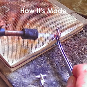 May include: A close-up of a metal piece being heated with a torch. The metal piece is held in place with tweezers on a ceramic tile. The text "How It's Made" is visible at the top of the image.