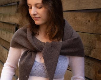 May include: A woman wearing a brown scarf tied around the neck, over a sheer white long-sleeved shirt and black jeans. The scarf is a dark brown color and has a triangular shape at the ends.