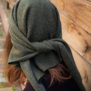 Puede incluir: Un gorro de punto verde oscuro, envuelto y atado en la parte delantera. El gorro está hecho de un material suave y texturizado. La persona lleva una camiseta negra. El fondo es una valla de madera.