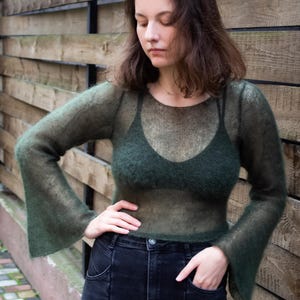 May include: A sheer, olive green long-sleeved top with flared sleeves. The top is paired with a dark green bra and black jeans. The model is standing in front of a wooden fence.