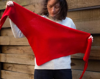May include: A vibrant red triangular shawl with long, pointed ends. The shawl is held up, showcasing its shape and color. The background is a wooden fence.