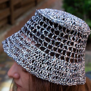 May include: A silver crocheted bucket hat with a wide brim. The hat is made of a metallic yarn, creating a shimmering effect. The open weave design allows for ventilation and a lightweight feel. The hat is a stylish accessory.