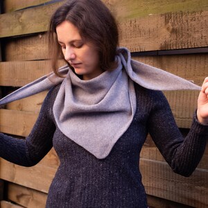 Puede incluir: Una mujer lleva una bufanda gris de forma triangular. La bufanda está hecha de un tejido suave y de punto y está drapeada alrededor de su cuello. Lleva un jersey azul oscuro y está de pie frente a una pared de madera.