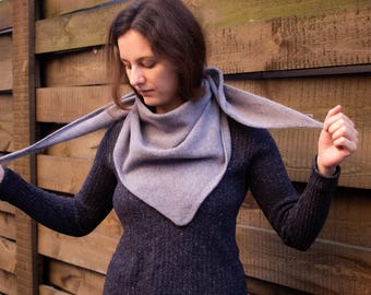 May include: A woman wearing a gray scarf with a triangular shape. The scarf is made of a soft, knit fabric and is draped around her neck. She is wearing a dark blue sweater and is standing in front of a wooden wall.
