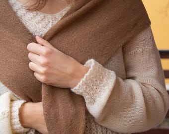 May include: A woman wearing a white knitted sweater with a brown knitted scarf draped around her neck. The scarf is tied in a knot at the front of her chest. She is wearing blue jeans and a brown leather belt.
