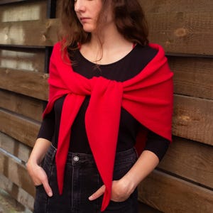 May include: A woman wearing a red shawl tied around the shoulders over a black long-sleeve shirt. She is wearing black jeans and standing in front of a wooden fence.